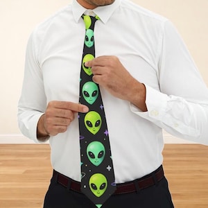 May include: A black necktie featuring a repeating pattern of green alien faces with large black eyes. The aliens are set against a black background with small white stars and purple accents. The tie is being worn with a white dress shirt.
