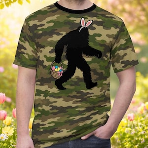 May include: Camouflage-patterned t-shirt featuring a black silhouette of Bigfoot wearing bunny ears and carrying a basket of Easter eggs. The shirt has a black collar and a short-sleeve design. The background is a blurred image of a garden.