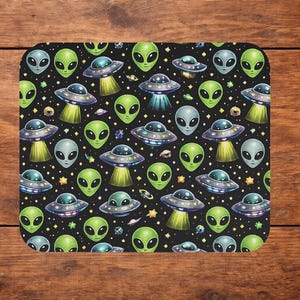 May include: A rectangular mouse pad featuring a repeating pattern on a black background. The design includes green alien faces with large black eyes and silver flying saucers emitting yellow light beams, interspersed with small yellow stars.