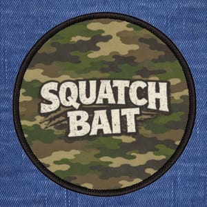 May include: Round patch with a camouflage pattern in shades of green, brown, and tan. The patch features the words "SQUATCH BAIT" in a distressed white font, outlined in black. The patch has a black border.