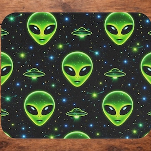 May include: A black mousepad featuring a repeating pattern of green alien faces and flying saucers against a starry background. The aliens have large black eyes and a smiling expression. The mousepad has rounded corners.