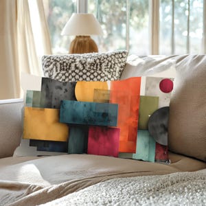 May include: Decorative throw pillow with an abstract design. The pillow features overlapping rectangular and circular shapes in various colors, including yellow, orange, teal, and red. The pillow is placed on a beige sofa.