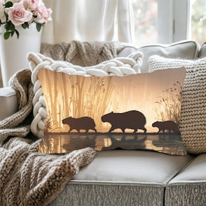 Puede incluir: Cojín decorativo rectangular con la silueta de tres carpinchos caminando por una masa de agua, con hierba alta en el fondo. El cojín tiene una cálida combinación de colores dorados, que evoca una escena de puesta de sol. El cojín está colocado en un sofá.