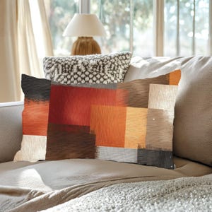 May include: Rectangular decorative pillow with an abstract design. The pillow features a patchwork of colors including black, red, orange, and beige. The pillow is placed on a neutral-toned sofa, creating a cozy and inviting atmosphere.