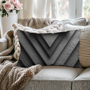 May include: Rectangular decorative pillow with a chevron pattern in shades of gray. The pillow has a textured appearance, and is placed on a light-colored sofa. The pillow is approximately 12 inches wide and 20 inches long.