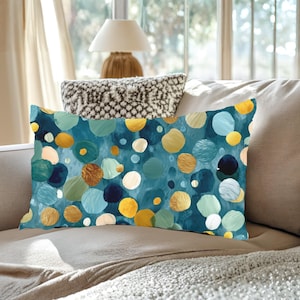 May include: Rectangular decorative pillow with a teal background and a pattern of colorful, overlapping circles in shades of gold, yellow, blue, and white. The pillow is placed on a neutral-toned sofa.