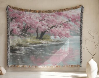 May include: A woven throw blanket depicting a tranquil scene of pink cherry blossom trees reflected in water. The artwork has a soft, painterly style with a fringed border. The colour palette includes shades of pink, grey, and green.