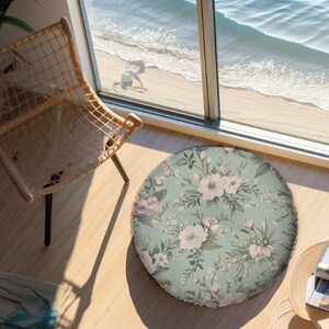 May include: A round floor cushion featuring a sage green background with a repeating botanical pattern of white and soft pink flowers, green leaves, and small buds. The cushion has a flat, circular shape and a smooth, fabric-covered surface.