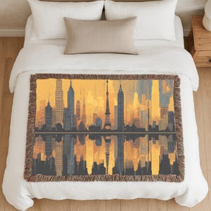 May include: A woven throw blanket featuring a cityscape design with a skyline of buildings in shades of yellow, gray, and blue. The design includes a reflection of the skyline below. The blanket has a fringed edge and is displayed on a bed.
