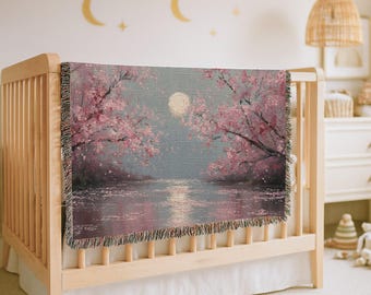 May include: A woven throw blanket featuring a landscape scene with pink cherry blossom trees on either side of a calm body of water under a full moon. The blanket has a textured, fringed edge and displays a soft colour palette of pink, grey, and white.