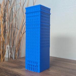 May include: A vibrant blue, miniature skyscraper model with detailed architectural features. The building is rectangular with multiple stories, windows, and decorative elements. It stands on a wooden surface, showcasing a modern design.