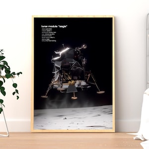 Op de afbeelding: Een ingelijste print met de maanlander "Eagle" op de maan. Het ruimtevaartuig is gedetailleerd afgebeeld, met uitgestrekte poten en zichtbare uitlaatgassen. De tekst bovenaan staat tegen een zwarte achtergrond, met een houten lijst.