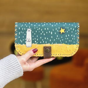 May include: A rectangular wallet with a whimsical design. The top portion is teal with white dots and a cartoon character and star. The bottom is yellow. A brown leather strap with a button secures the wallet.