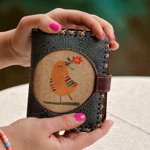May include: A dark green leather wallet with a stitched border and a brown leather strap. The front features a circular design with an orange bird holding a red flower. The bird has colorful striped details.