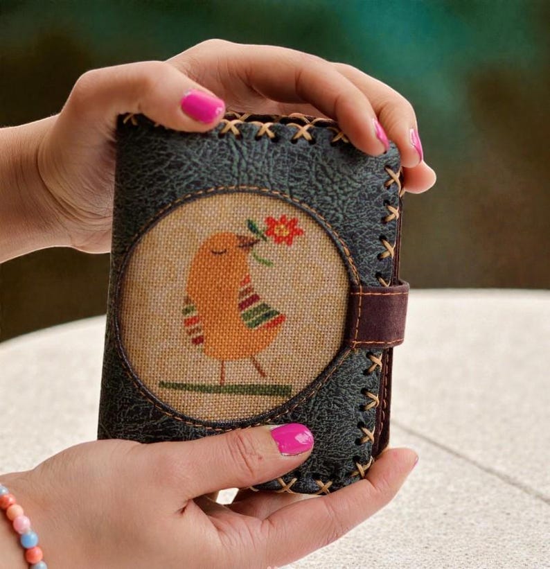 May include: A dark green leather wallet with a stitched border and a brown leather strap. The front features a circular design with an orange bird holding a red flower. The bird has colorful striped details.