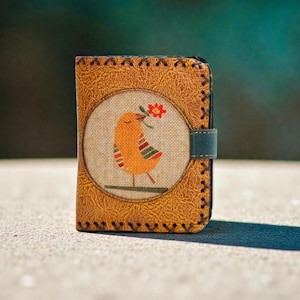 Könnte beinhalten: Eine handgefertigte Ledergeldbörse mit einer genähten Bordüre und einem Vogeldesign. Der orangefarbene Vogel mit roten und grünen Akzenten hält eine rote Blume. Die Geldbörse hat einen teal Verschluss und eine strukturierte, hellbraune Lederaußenseite.