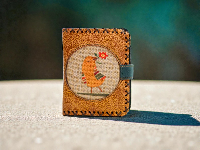May include: A handcrafted leather wallet with a stitched border and a bird design. The bird is orange with red and green accents, holding a red flower. The wallet has a teal clasp and a textured, tan leather exterior.
