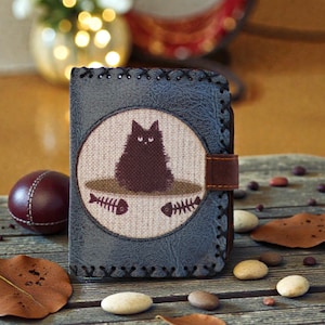 May include: A blue leather wallet with a stitched border and a brown leather strap closure. The front features a circular patch with a black cat illustration above two fish skeletons. The wallet is on a wooden surface.