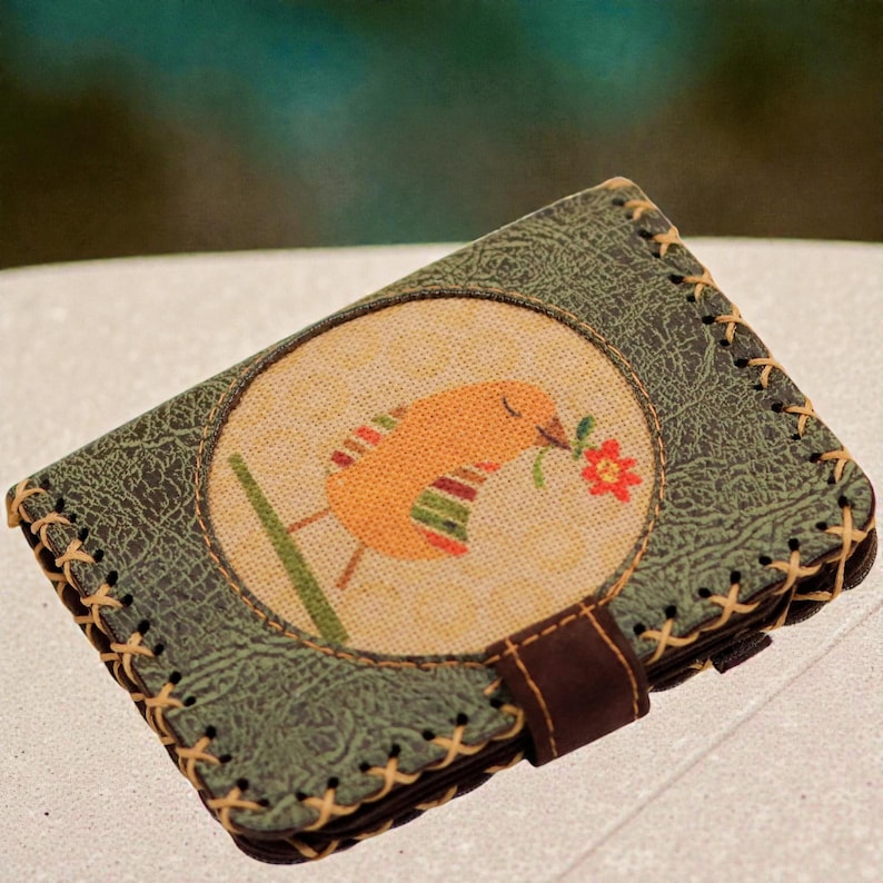 May include: A green leather wallet with a stitched design and a round embroidered patch featuring a yellow bird holding a red flower. The wallet has a brown strap closure.