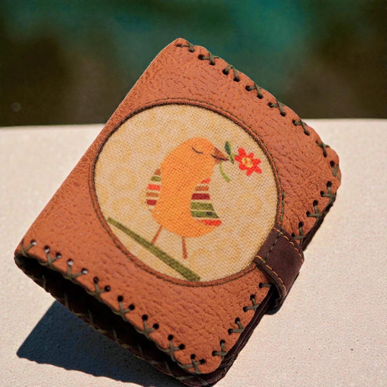 May include: Brown leather wallet with a stitched border. The front features a circular design with an orange bird holding a red flower. The wallet has a dark brown strap closure.