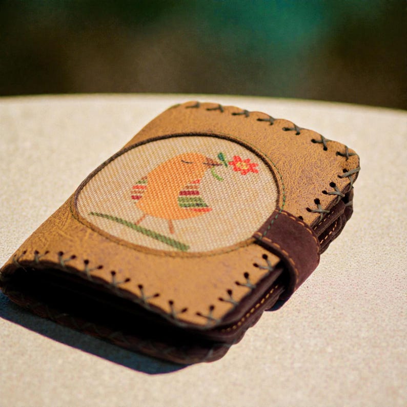 May include: A brown leather wallet with a stitched border and a circular fabric patch. The patch features an orange bird holding a red flower. A dark brown strap secures the wallet closed. The wallet is made of leather.