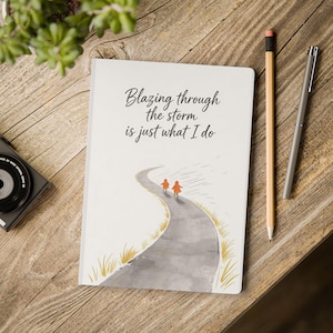 Puede incluir: Un cuaderno blanco con el texto "Blazing through the storm is just what I do" y una ilustración de dos figuras caminando por un camino. Un lápiz y un bolígrafo sobre una superficie de madera.