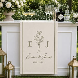 May include: A cream-colored fabric table runner featuring a dark green line-art floral illustration, the letters E and J, the names Emma & James, and the date 27.07.2027. The runner is decorated with two vertical lines along the side edges.