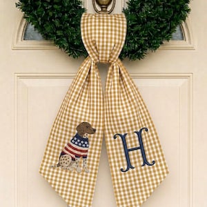 May include: A mustard yellow and white gingham fabric door hanger featuring an embroidered brown dog wearing a red, white, and blue striped sweater on the left side and a large navy blue embroidered letter H on the right side.