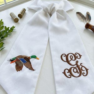 May include: A white fabric necktie featuring an embroidered mallard duck in flight on one side and a brown monogrammed letter G in a decorative script font on the other side. The tie is knotted at the top and made of a smooth, solid white material.