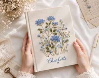 Può includere: Un libro rivestito in tessuto color crema con un intricato ricamo di fiori selvatici blu e bianchi con steli verdi. Il nome Charlotte è ricamato in corsivo blu sotto il disegno floreale, accompagnato da un piccolo cuore blu.