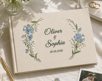 Può includere: Un libro degli ospiti rettangolare rivestito in tessuto color crema, decorato con rametti di fiori ricamati in blu e bianco su entrambi i lati. Al centro presenta il testo Oliver & Sophia e la data 26.06.2026 in un carattere corsivo e serif verde scuro.