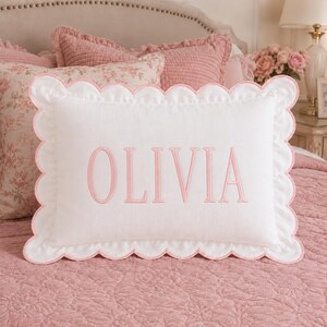 May include: A rectangular white pillow featuring a scalloped edge trimmed in light pink. The center of the pillow displays the name OLIVIA embroidered in a light pink serif font. The pillow has a textured fabric finish and a clean design.