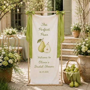 May include: A vertical cream-colored fabric banner suspended on a wooden frame, featuring green satin ribbons at the top corners. The text reads The Perfect Pear, Welcome to Olivia's Bridal Shower, and 06.25.2026, with a pear illustration.