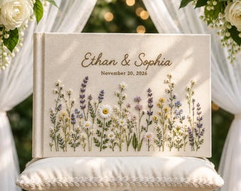 Può includere: Un album fotografico rettangolare in tessuto color crema con fiori di campo ricamati in tonalità viola, gialle e bianche. La copertina riporta il testo Ethan & Sophia e la data November 20, 2026, in un elegante carattere marrone.