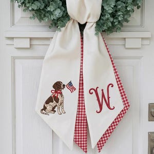 May include: A cream-colored fabric door hanger featuring a green eucalyptus wreath at the top. The left panel displays an embroidered brown and white spotted dog with a red bow and an American flag. The right panel features a red embroidered letter W.