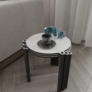 Puede incluir: Una mesa auxiliar moderna con tapa blanca y patas negras. La mesa presenta un diseño decorativo con una taza y un platillo, y dos figuras de elefantes azules. La mesa está colocada junto a un sofá de color claro y una cortina transparente.