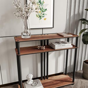 Puede incluir: Una consola moderna de tres niveles con acabado en madera oscura y soportes de metal negro. El estante superior contiene un libro y una bandeja, el estante central tiene esferas decorativas y el estante inferior muestra un busto y libros. Una impresión botánica enmarcada cuelga encima.