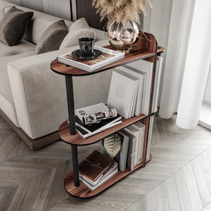 Peut inclure: Une table d'appoint à trois niveaux en bois avec des supports en métal noir. L'étagère supérieure contient une tasse et une soucoupe, un livre et un vase décoratif. Les étagères inférieures présentent des livres et un journal en cuir. La table est placée devant un fond neutre.