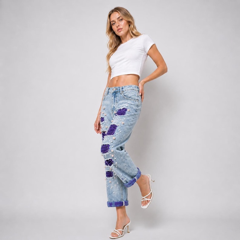 May include: Light blue denim jeans with distressed details and purple sequin patches. The jeans are embellished with faux pearls and have cuffed hems. The model is wearing a white crop top and white heels.