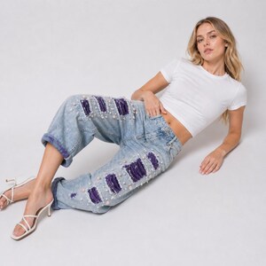 May include: Light blue denim jeans with distressed patches and pearl embellishments. The jeans have a relaxed fit and are cuffed at the ankles. The model is wearing a white t-shirt and white heels.