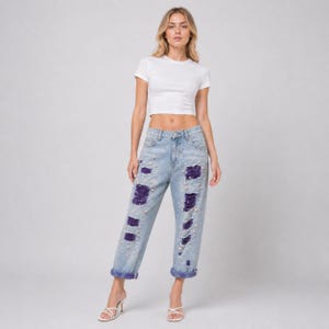 May include: Light wash denim jeans with distressed patches and pearl accents. The jeans have a relaxed fit and are cuffed at the ankles. The patches are a deep purple color. The model is wearing a white crop top.