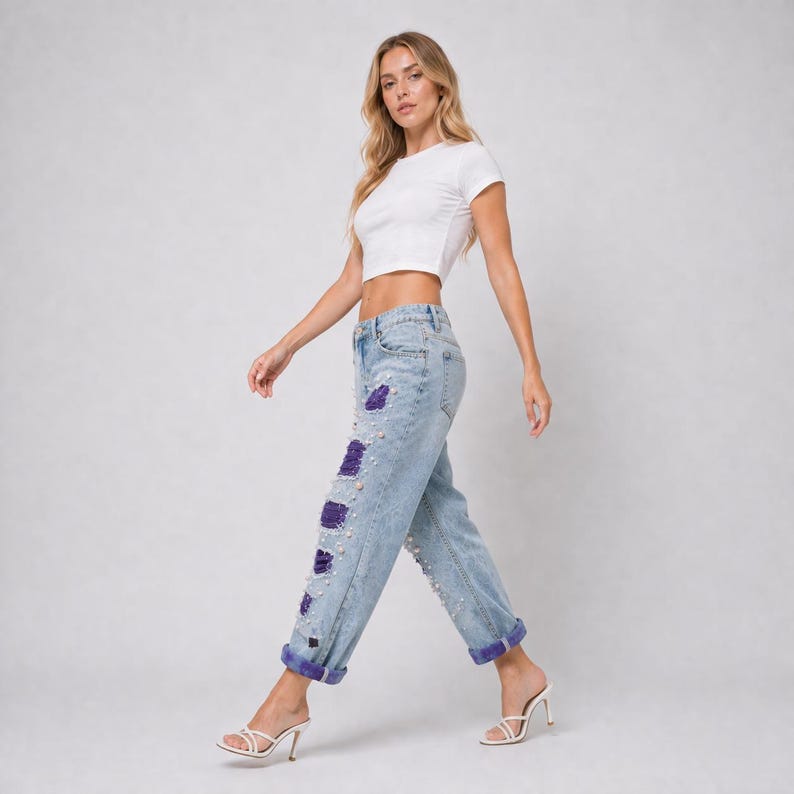 May include: Light wash denim jeans with distressed patches, embellished with purple fabric and faux pearls. The jeans have a relaxed fit, cuffed hems, and are paired with a white crop top.