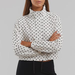 May include: A white cropped jacket with a black polka dot pattern. The jacket has a high collar, long sleeves, and a drawstring at the waist. The model is wearing black athletic pants.