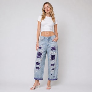 May include: Light blue denim jeans with distressed detailing, featuring ripped sections with dark purple fabric underneath. The jeans are embellished with small, pearl-like beads. The model is wearing a white crop top.