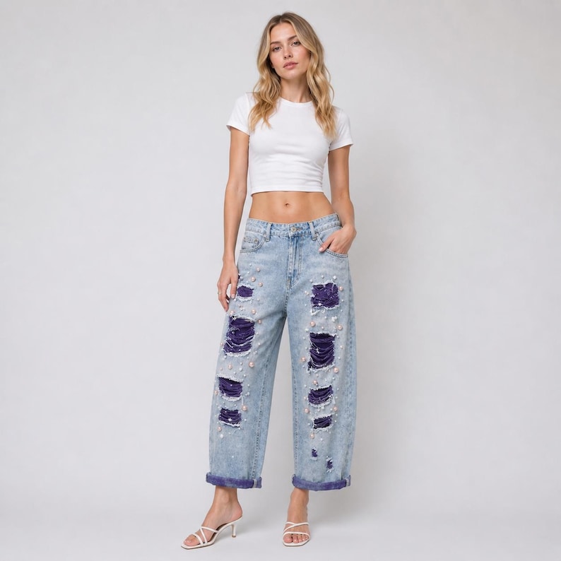 May include: Light blue denim jeans with distressed detailing, featuring ripped sections with dark purple fabric underneath. The jeans are embellished with small, pearl-like beads. The model is wearing a white crop top.