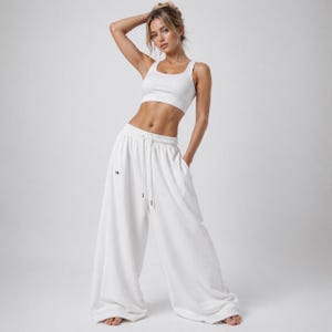 Könnte beinhalten: Ein weißes Sportoutfit, bestehend aus einem Crop-Top und einer weit geschnittenen Jogginghose. Das Crop-Top hat einen Rundhalsausschnitt. Die Jogginghose hat einen Kordelzug in der Taille und ein kleines Logo am linken Bein.