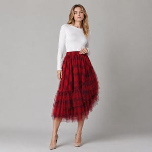 May include: A woman wearing a white long-sleeve top and a red plaid tulle skirt. The skirt has multiple layers and a ruffled hem. The outfit is set against a solid gray background.