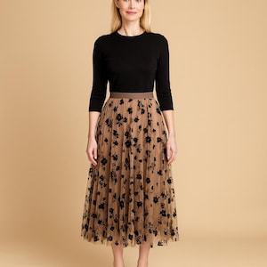 Puede incluir: Una mujer con una blusa negra de manga tres cuartos y una falda de tul color beige. La falda está adornada con bordados florales negros. Este atuendo es adecuado para diversas ocasiones, combinando elegancia y comodidad.