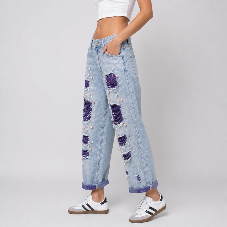 May include: Light blue denim jeans with distressed detailing, featuring purple patches and pearl embellishments. The jeans have a relaxed fit and are cuffed at the ankles.