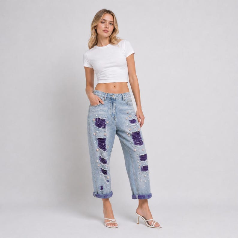 May include: A woman wearing a white crop top and light blue denim jeans. The jeans have distressed details with purple patches and pearl embellishments. The jeans are cuffed at the ankles.